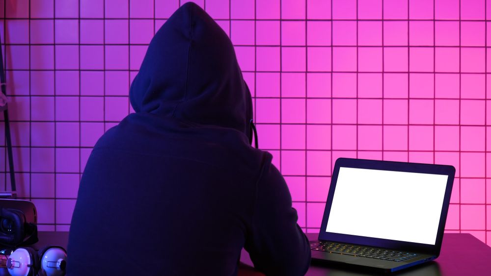 A person wearing a hoodie sits at a desk, focused on their laptop while working or studying.