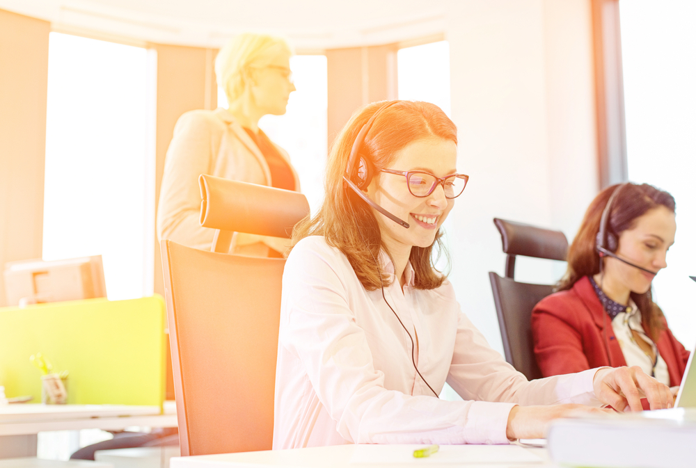 A woman in a headset at a desk, accompanied by two other women, focused on a collaborative task in an office.