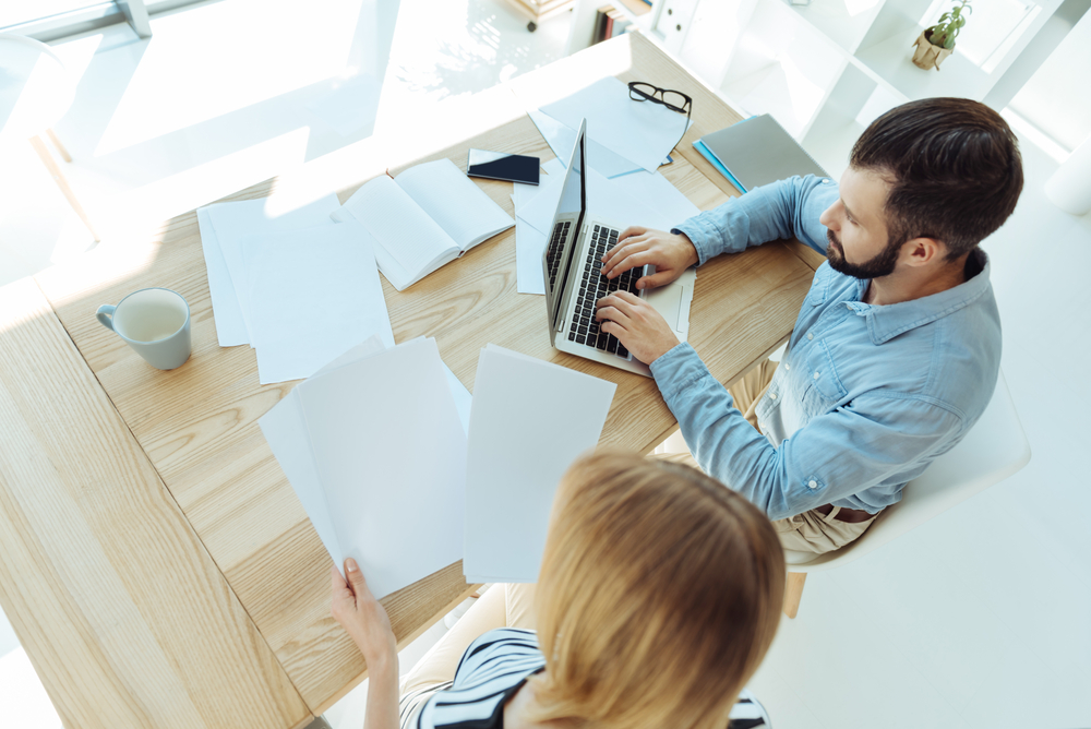 A man and woman sit at a table, reviewing documents and discussing details together.