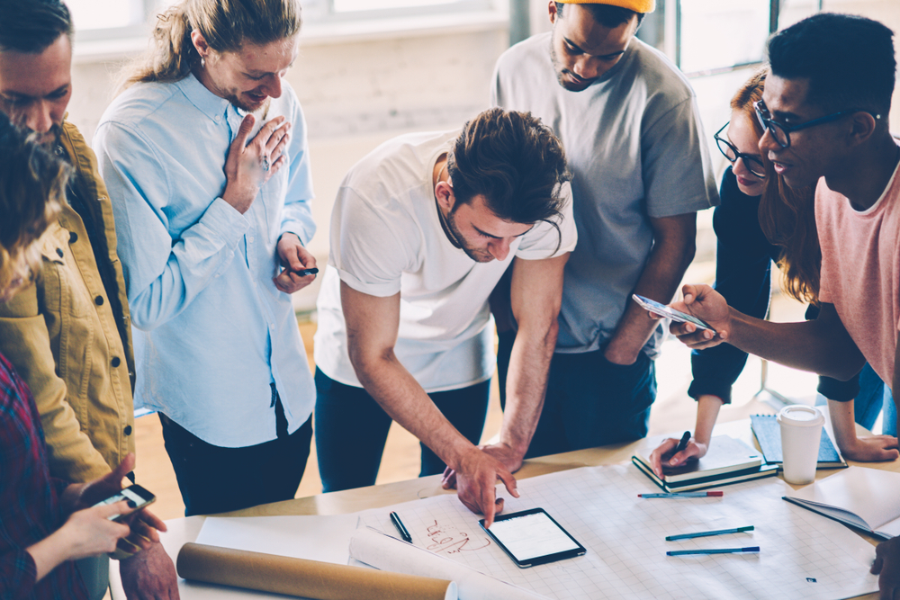 A diverse group of people surrounding a table, interacting with a tablet during a meeting or brainstorming session.