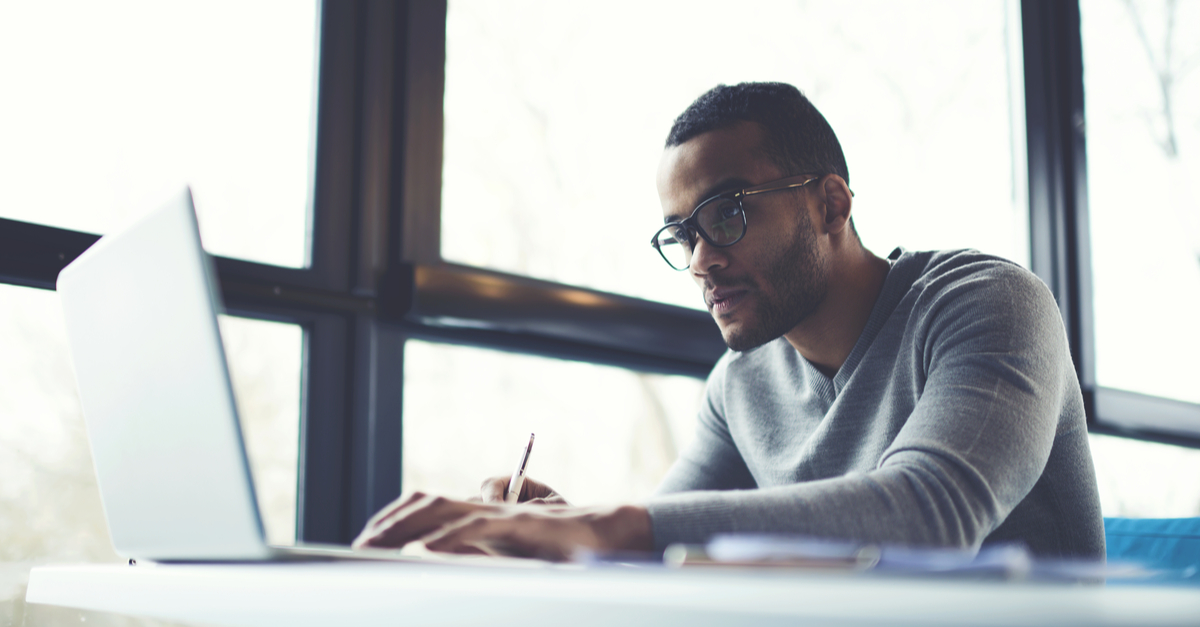 A man wearing glasses is focused on working on his laptop at a desk.
