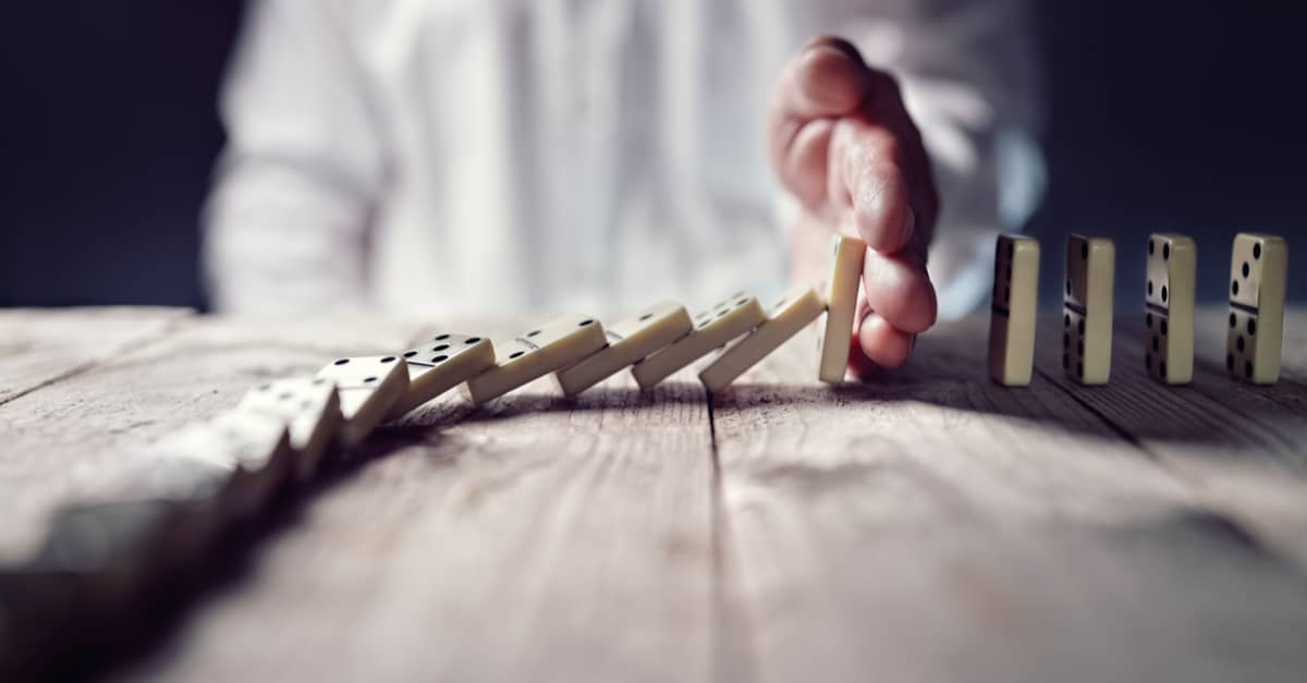 A person carefully places hand on a wooden table, stopping the chain reaction of falling dominoes.