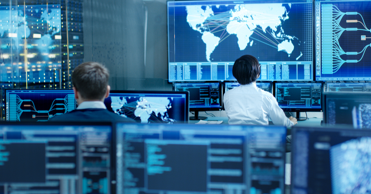 Two people are positioned before multiple computer screens, likely working together on a project or task.