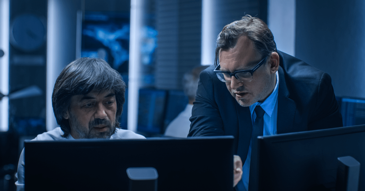 Two suited men examining data on computer screens, indicating a collaborative work environment.