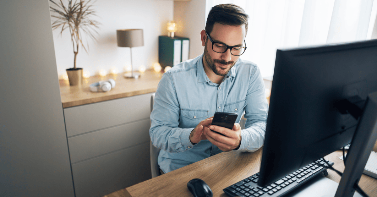 A man wearing glasses sits at a desk, focused on his phone while working.