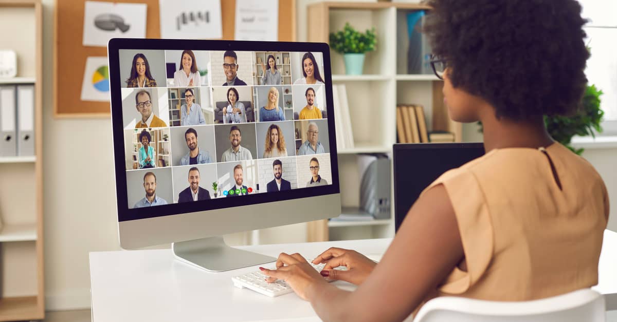 A woman sits at a desk, focused on a computer screen displaying a video call with a group of people.