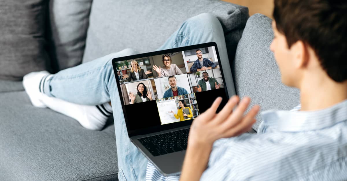 A man sits on a couch with a laptop, engaging in a video call with a group of people displayed on the screen.