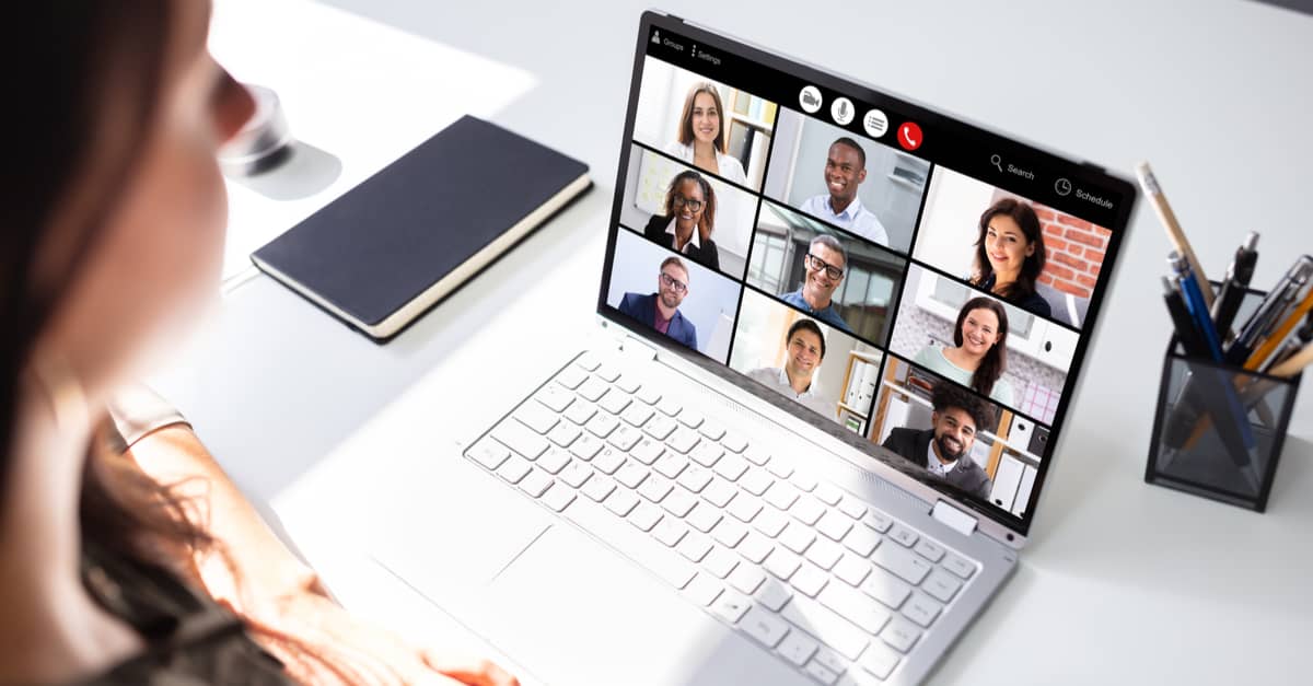 A woman sits at a desk with a laptop displaying several people on the screen during a virtual meeting.