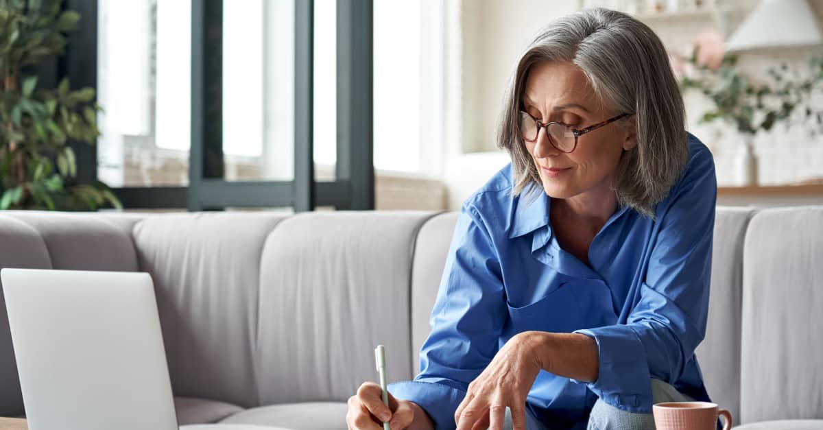 A woman wearing glasses is focused on writing on a piece of paper at a desk.