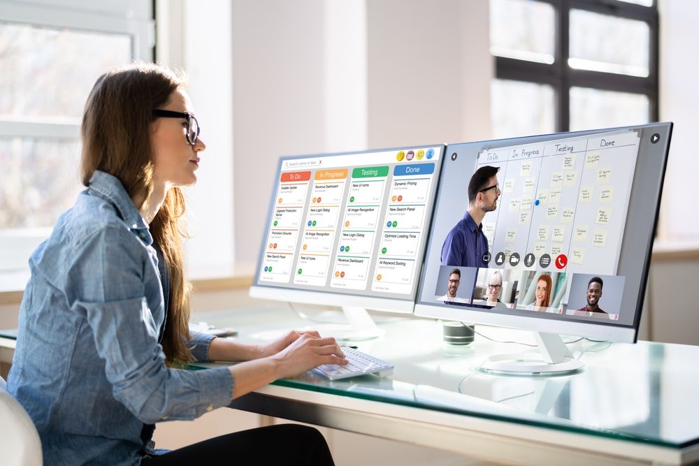 A woman seated at a desk, working on two computer monitors displaying various applications.