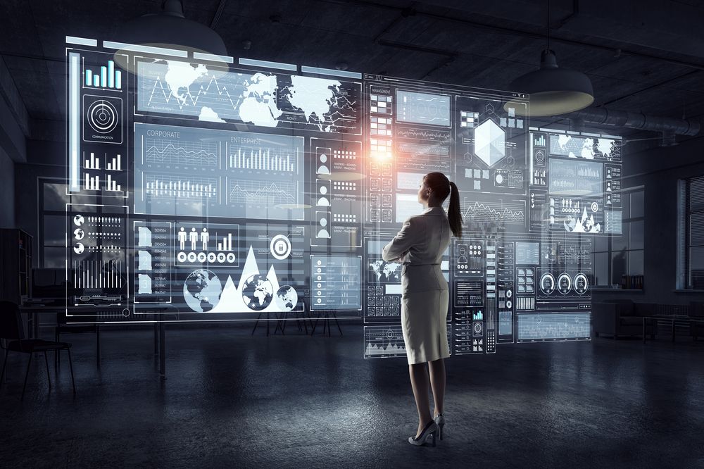 A woman in business attire stands before a large screen showing analytics information.